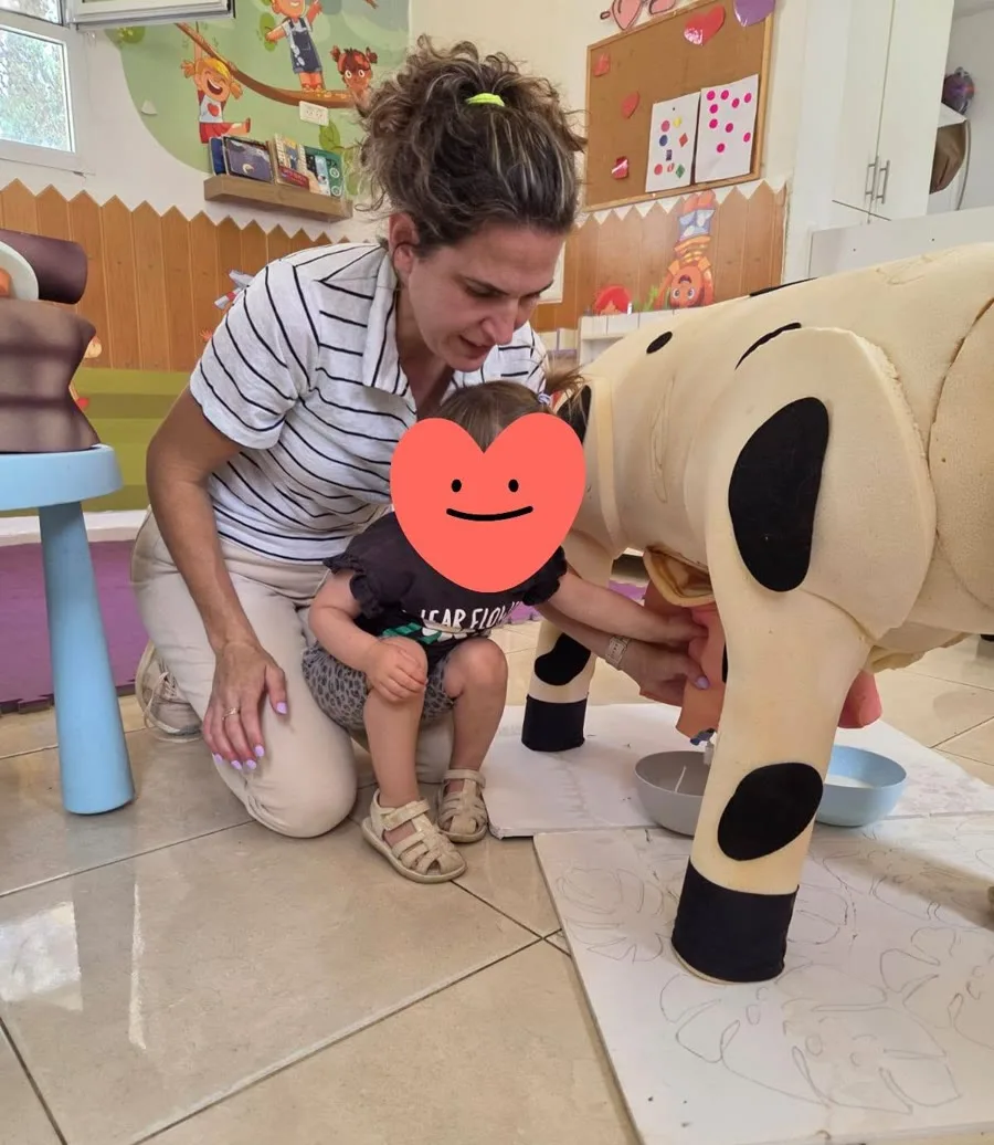 אשת צוות מלווה פעוטה בהדגמת חליבה על פרה מדומה בכיתת הגן